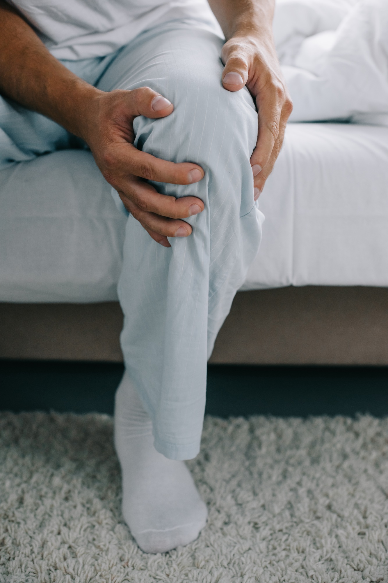 cropped shot of man in pajamas sitting on bed and suffering from pain in knee